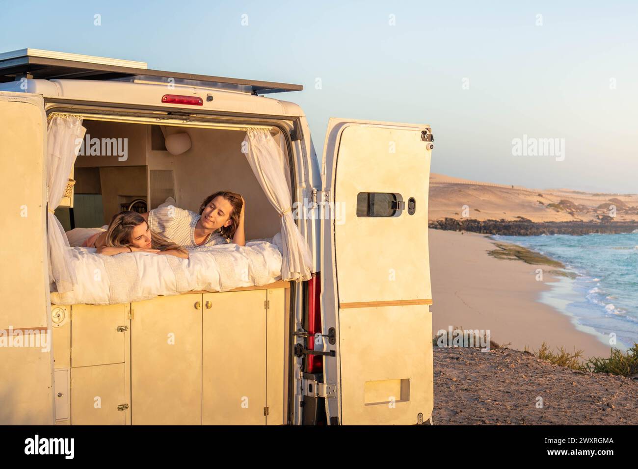 Ein paar Frauen werden entspannt auf dem Bett eines Wohnwagens gesehen, wie sie die aufgehende Sonne mit einem ruhigen Gesichtsausdruck betrachten. Stockfoto
