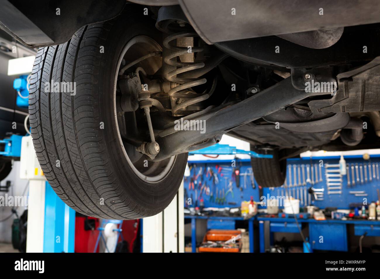 Ansicht von unten auf angehobenes Autorad mit Feder und Stoßdämpfer zur Reparatur in der Garage gegen ordentlich angeordnete mechanische Werkzeuge und Instrumente in Stockfoto