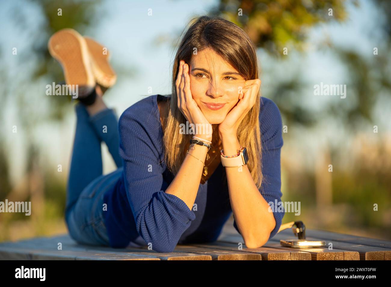Entspannte Frau, die mit nachdenklichem Gesicht wegschaut, während sie bei Sonnenuntergang auf einer Parkbank liegt. Stockfoto