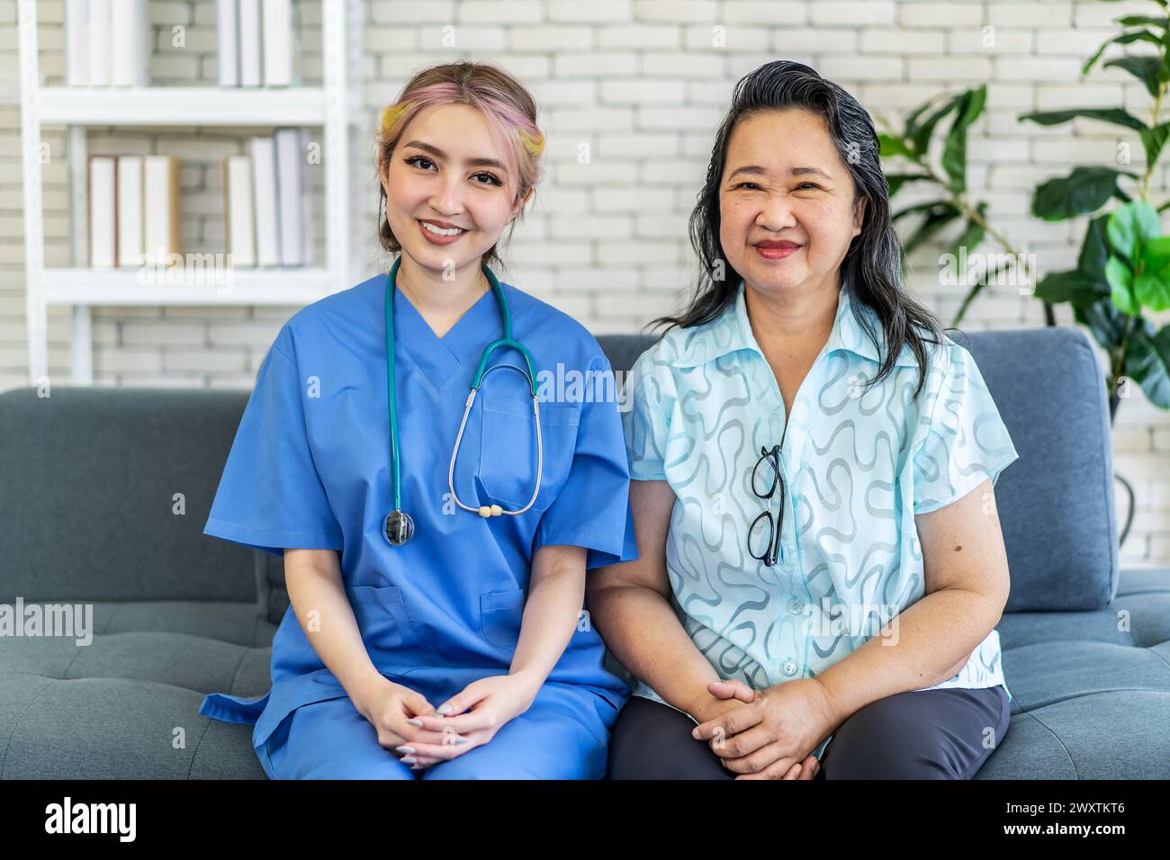 Eine Krankenschwester und eine asiatische Dame sitzen auf dem Sofa und schauen in die Kamera Stockfoto