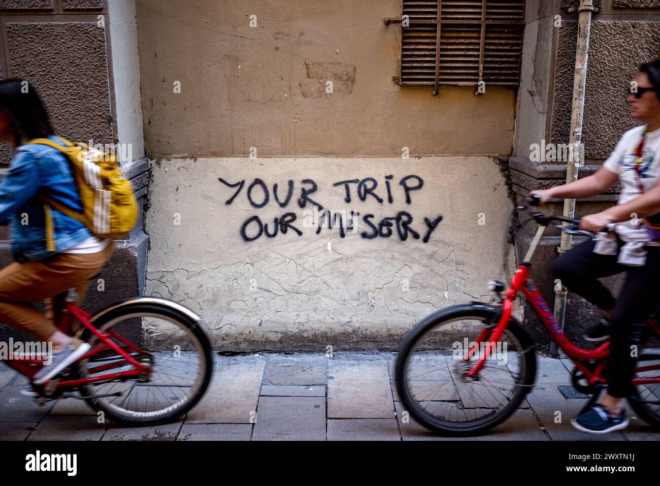 Im gotischen Viertel Barcelonas kommt eine Gruppe von Touristen auf einer Fahrradtour an einem Anti-Tourismus-Graffiti vorbei, das Ihre Reise unser Elend liest. In der katalanischen Hauptstadt gibt es seit Jahren Proteste gegen den Massentourismus. Katalonien hat eine Rekordzahl an Besuchern in den Osterferien 2024. Stockfoto