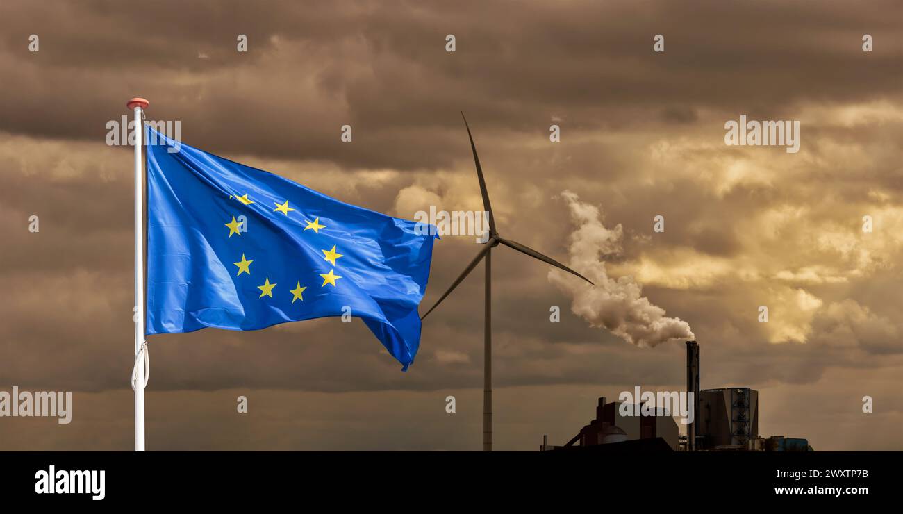 Europäische Flagge vor einem schadstoffbelastenden Fabrikschornstein mit Rauch- und Windkraftanlage schwenken Stockfoto