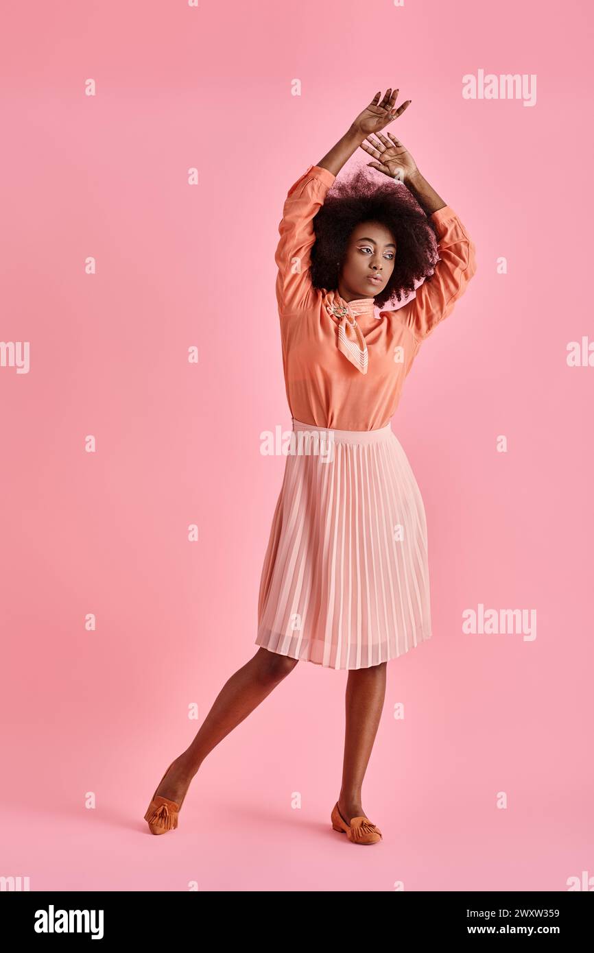 Lockige afroamerikanerin in Pfirsichbluse und Midirock posiert mit erhobener Hand auf Pastellrosa Stockfoto