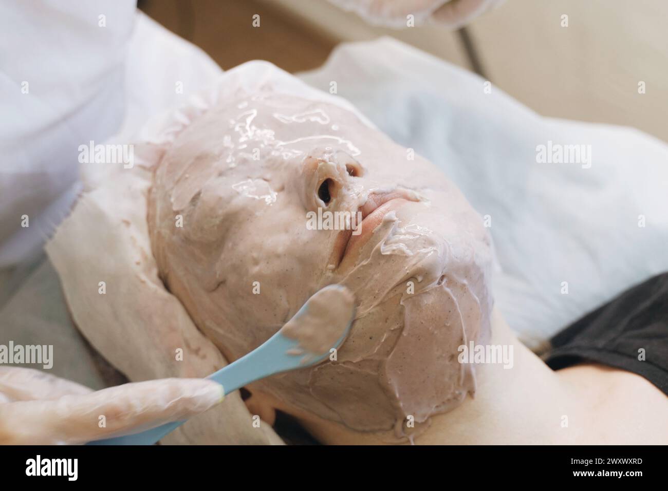 Eine Frau entspannt sich, während eine Spa-Therapeutin eine dicke Tonmaske auf ihr Gesicht aufträgt, die alles außer Augen und Mund für die Hautpflege bedeckt. Stockfoto