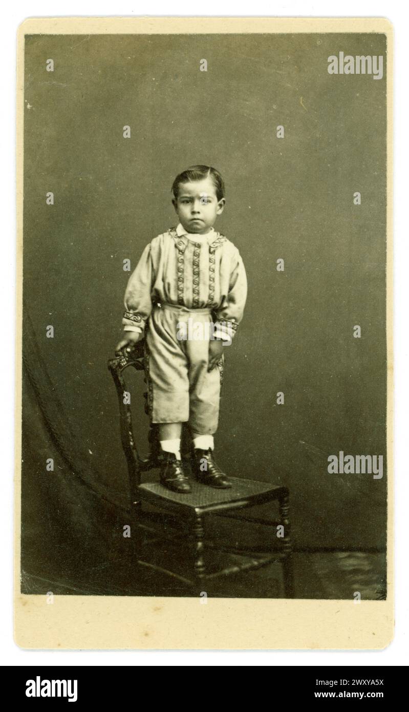 Original viktorianische Carte de Visite (Visitenkarte oder CDV) von süßem jungen viktorianischen Jungen, viktorianischem Kind, im Alter von etwa 3 oder 4 Jahren, mit einem aufwendigen Hemd, einem Hosenbein höher als die anderen, Stiefeln, die mürrisch aussehen, 1860er Jahre, Großbritannien Stockfoto