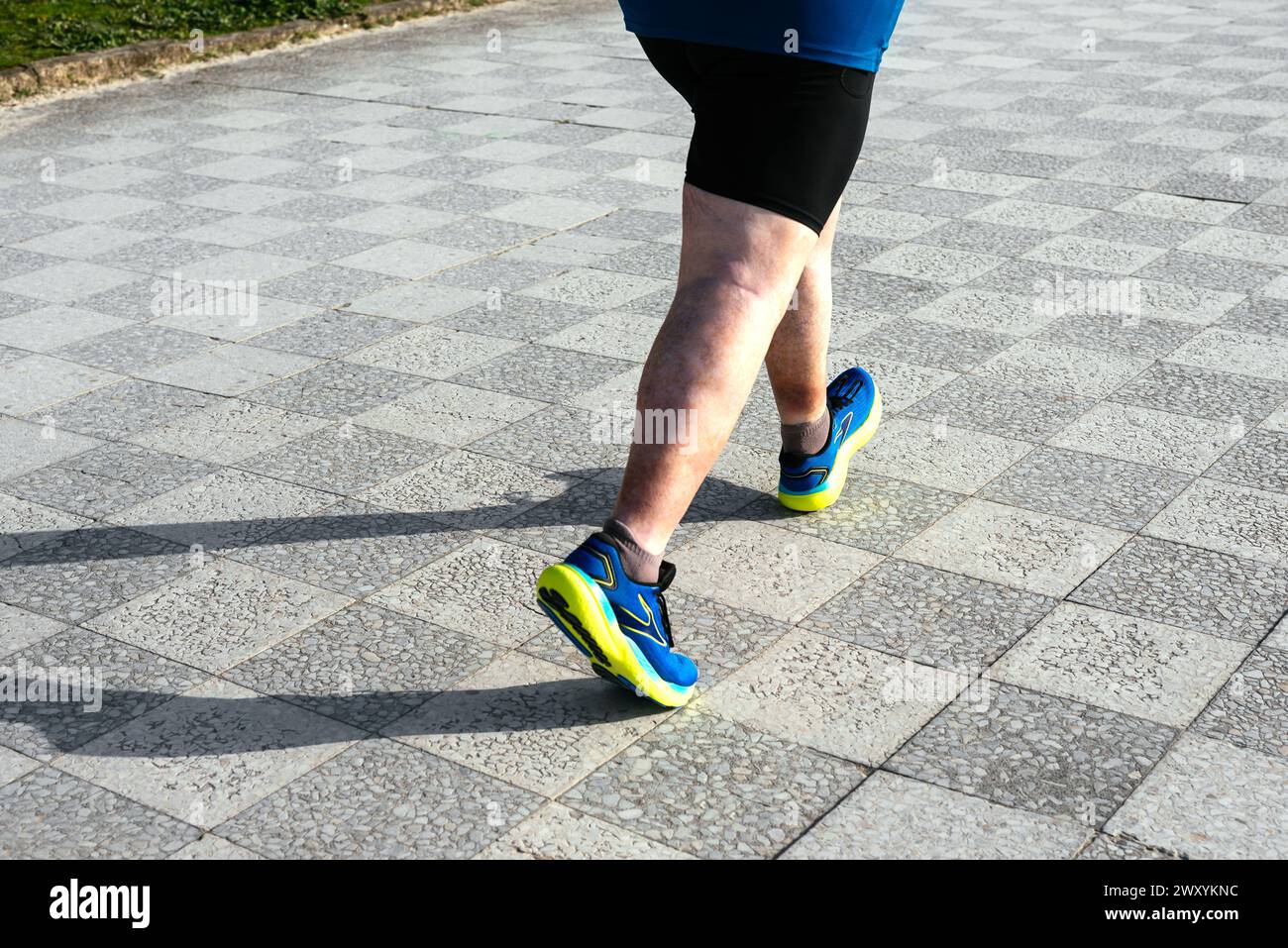 Der Unterkörper einer Person wird im mittleren Schritt auf einem gemusterten Bürgersteig mit sportlichen blauen und gelben Schuhen und schwarzen Shorts festgehalten Stockfoto