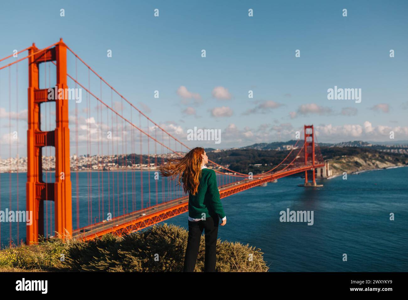 Rückansicht Frau steht in Ehrfurcht, während der Wind ihr Haar fängt und an einem lebhaften Frühlingstag die herrliche Golden Gate Bridge überblickt Stockfoto