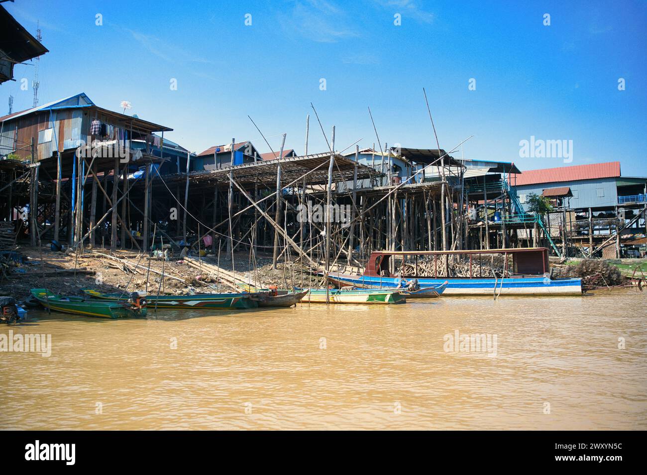 Hölzerne Stelzenhäuser, die sich über einem schlammigen Fluss erheben, mit traditionellen Booten, die am Ufer unter einem klaren blauen Himmel vertäut sind Stockfoto