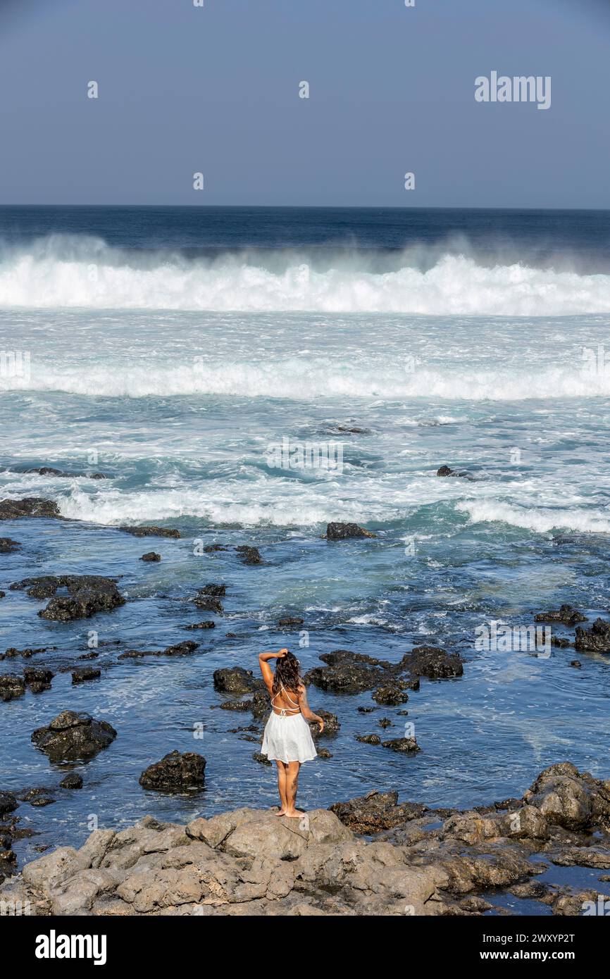 Ein Weibchen blickt über schaumige Wellen vom felsigen Ufer und verkörpert eine friedliche Wochenendreise-Atmosphäre. Stockfoto
