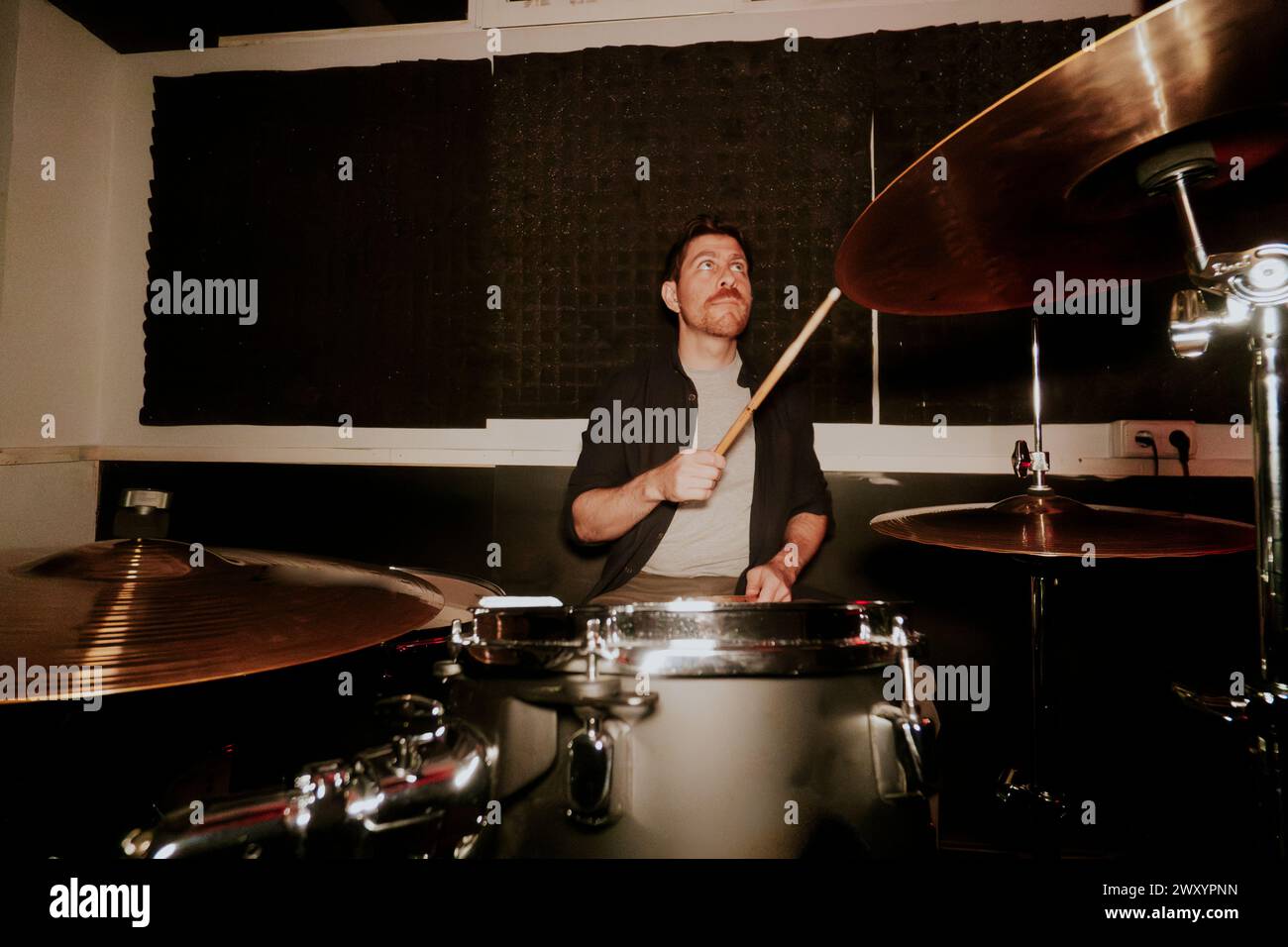 Ein fokussierter männlicher Schlagzeuger spielt Schlagzeug in einem schwach beleuchteten Studio und blickt von der Kamera weg. Stockfoto