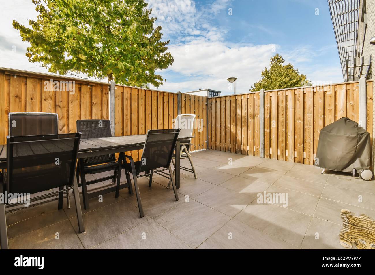 Ein einladender urbaner Hinterhof mit Esstisch, Stühlen und überdachtem Grill mit Holzzaun und reifem Baum. Stockfoto