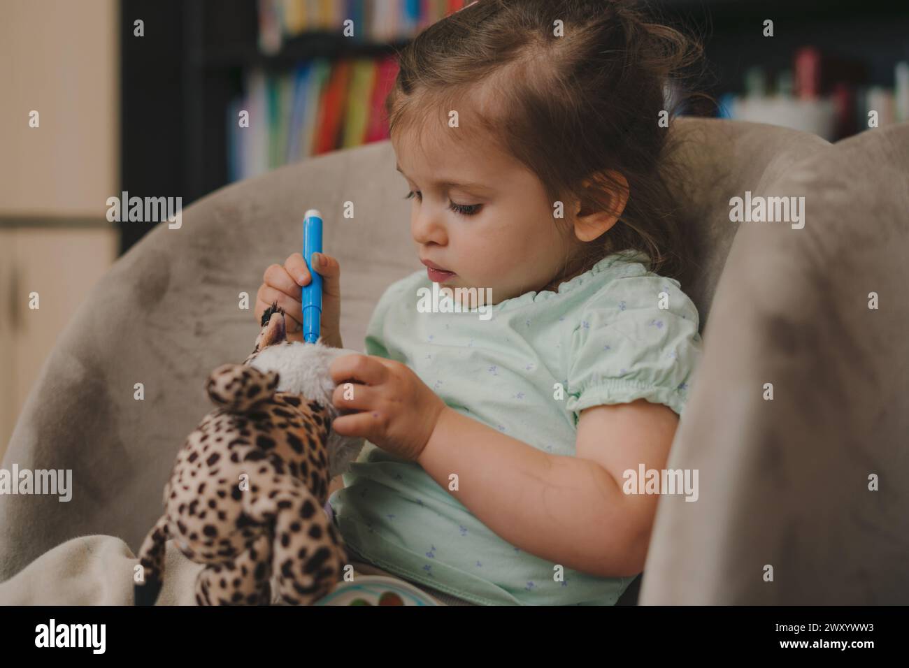 Porträt eines kleinen kaukasischen Mädchens, das zu Hause einen blauen Filzstift auf dem Schlafsofa hält und ein Kuscheltier verschmutzt. Kindheit Freizeit Spaß spielen Lernen Stockfoto
