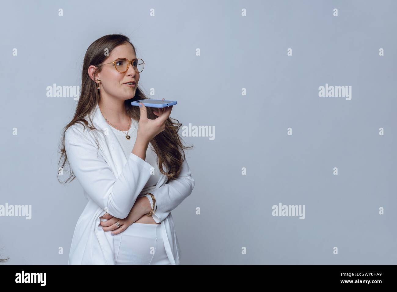Junge lateinfrau in weißer Kleidung, mit langen Haaren und Brillen, glückliche Geschäftsfrau, die steht und eine Sprachnachricht mit ihrem Telefon sendet, Studio-Sho Stockfoto