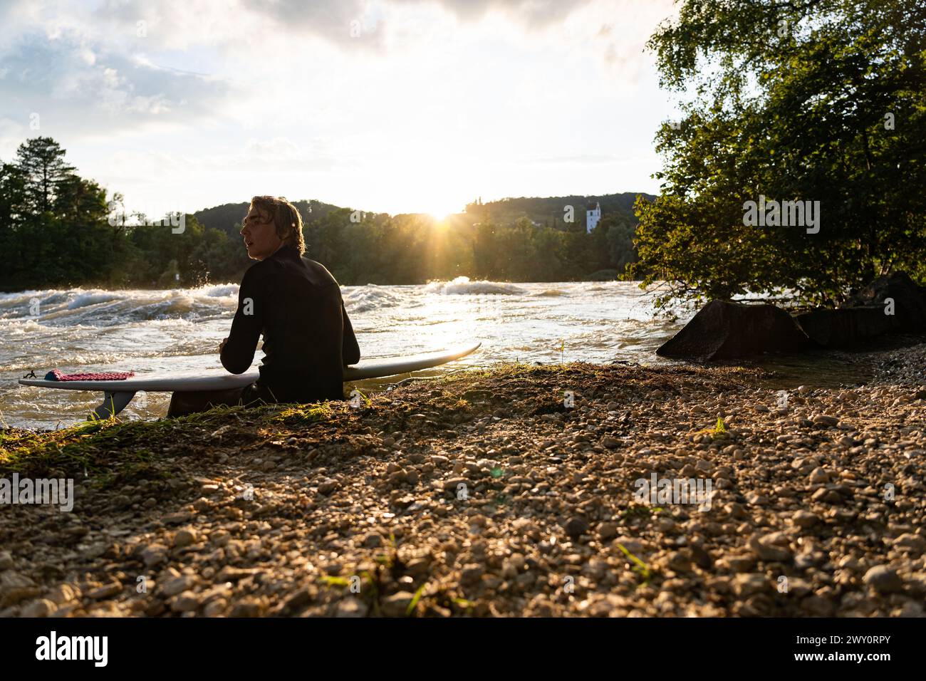 Der junge Surfer sitzt am Fluss Aare, schaut den Sonnenuntergang und genießt die ruhige Atmosphäre. Stockfoto