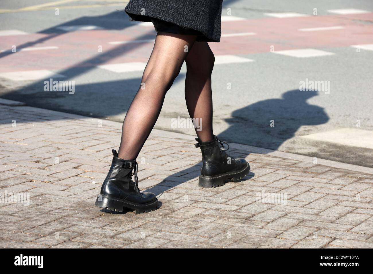 Weibliche Beine in schwarzen Stiefeln mit Schatten auf dem Bürgersteig. Schlankes Mädchen in Strumpfhosen, das eine Straße entlang läuft, Mode in der Frühlingsstadt Stockfoto