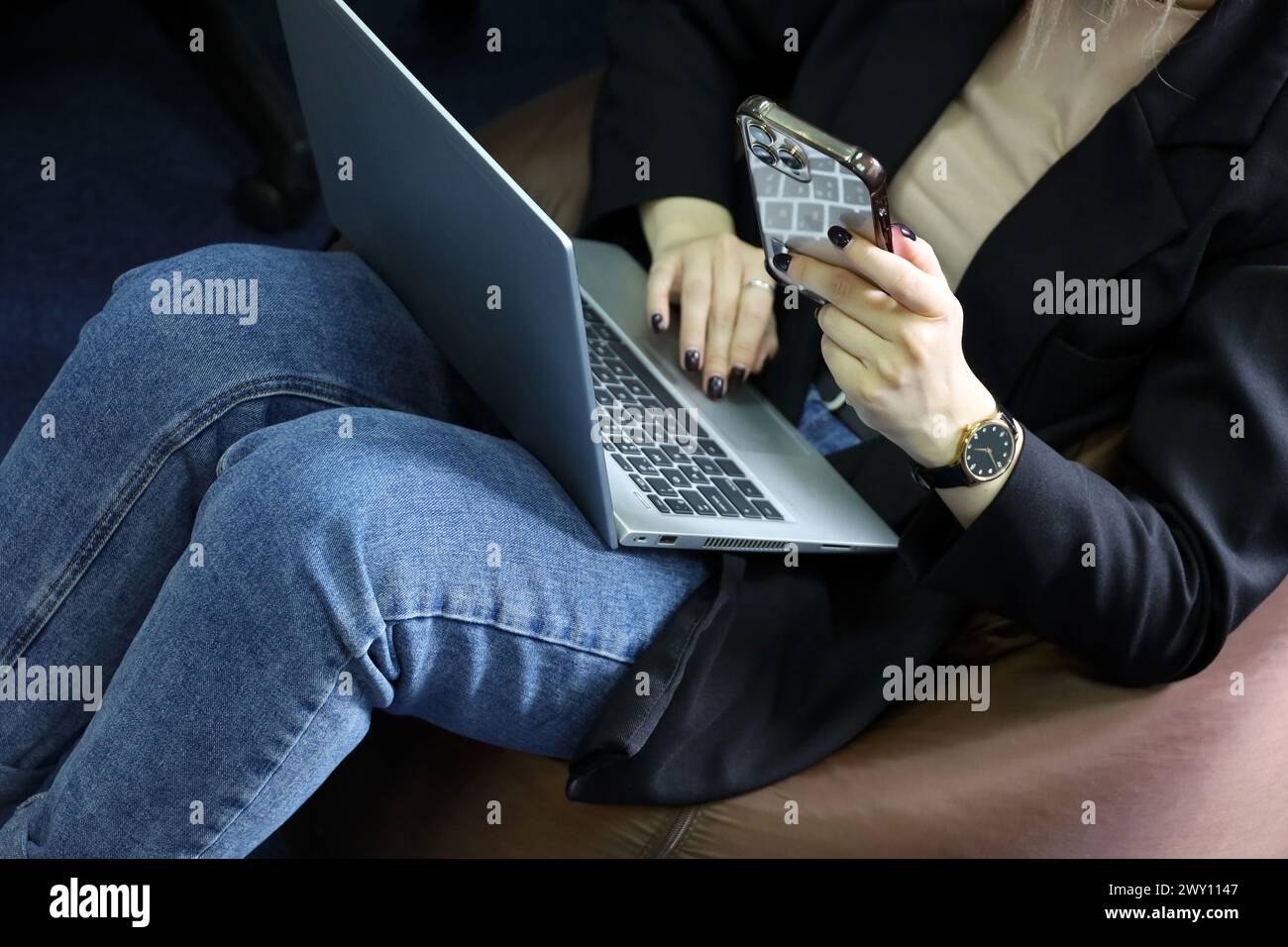 Mädchen in Jeans auf gepolstertem Hocker mit Laptop auf den Knien und Smartphone. Komfortables Arbeiten im Büro Stockfoto