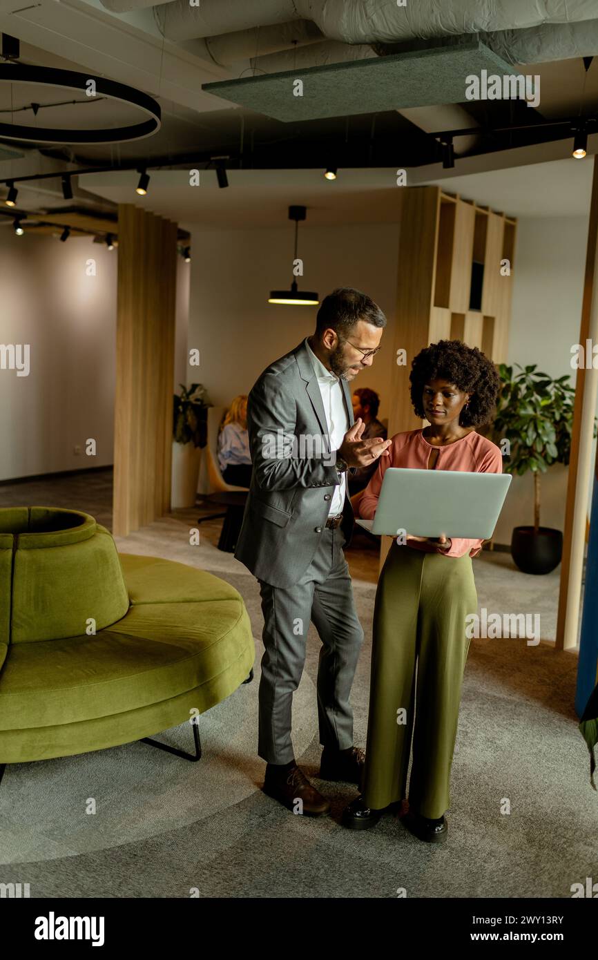 Mann und Frau diskutieren die Arbeit auf einem Laptop in einer lebhaften Büroumgebung, mit Kollegen im Hintergrund Stockfoto