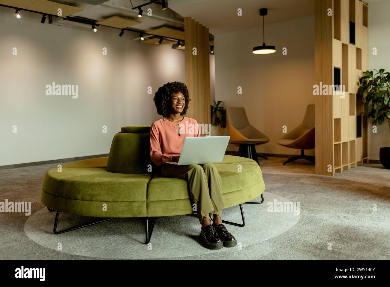 Eine temperamentvolle afroamerikanische Geschäftsfrau lächelt, während sie an ihrem Laptop arbeitet, bequem in einer lebendigen Bürolounge sitzt. Stockfoto