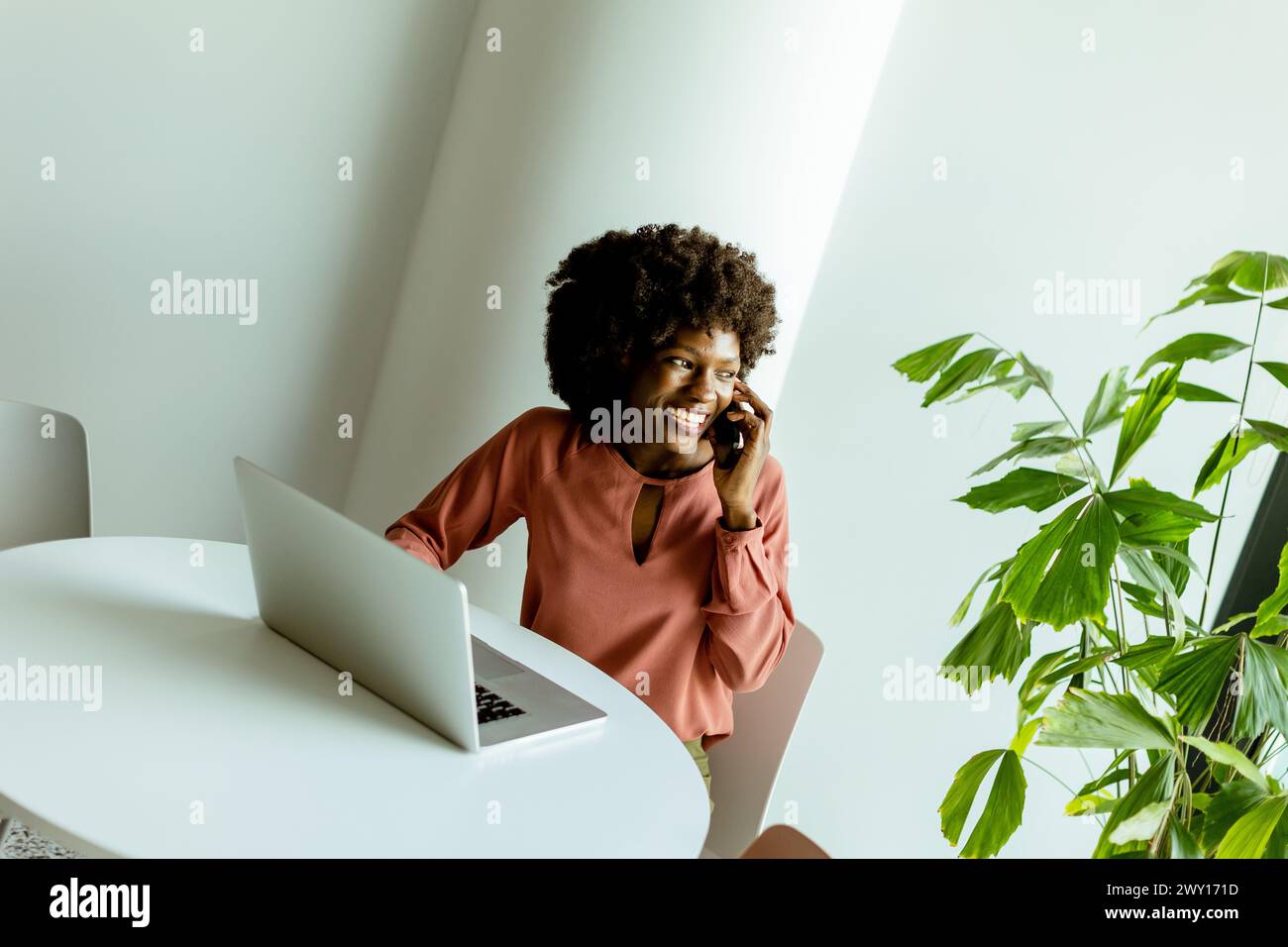 Eine fröhliche Frau spricht am Telefon an einem Café-Tisch mit einem Laptop und einer lebendigen Pflanze neben ihr. Stockfoto