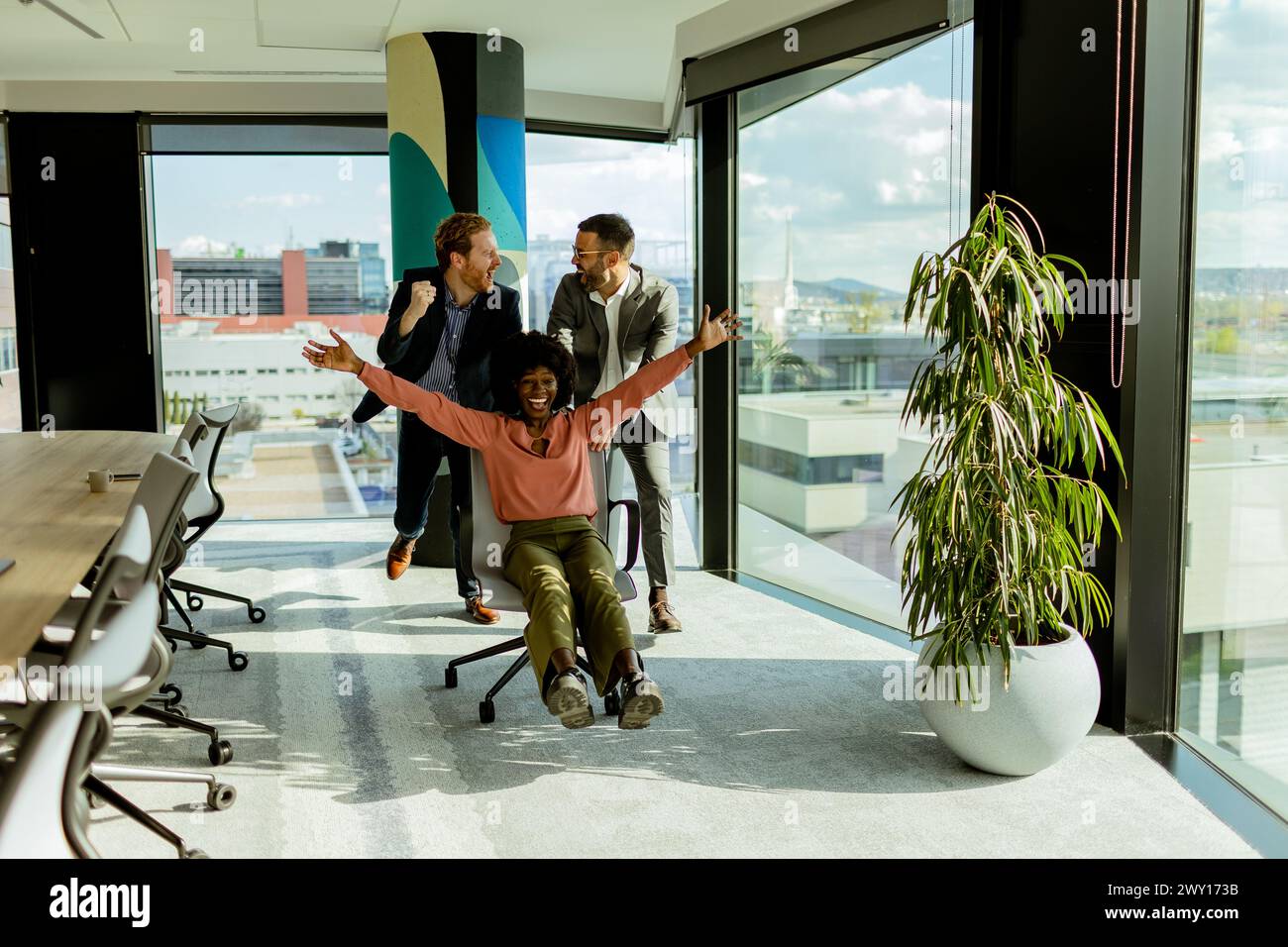 Überschwängliche Kollegen nehmen an einem spontanen Rennen mit Bürostühlen Teil, während sie sich im Nachmittagslicht vor Lachen sonnen. Stockfoto
