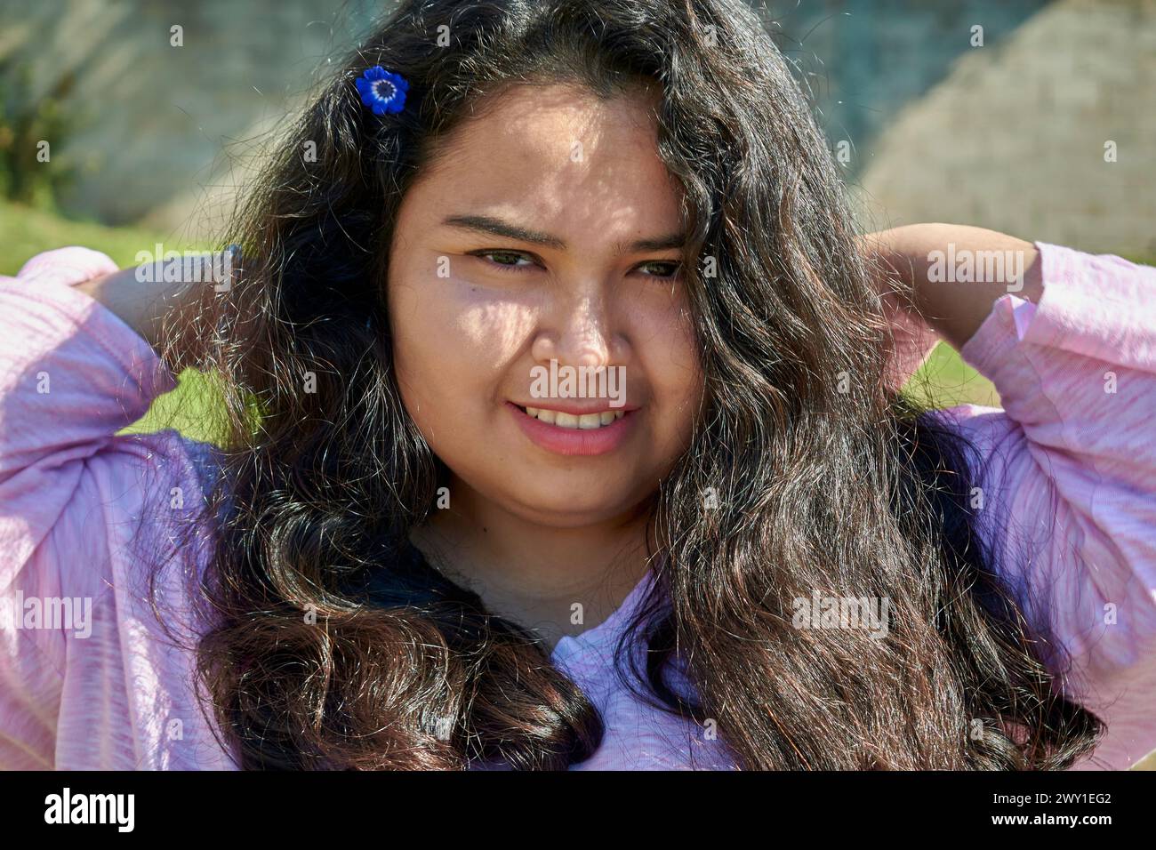 Porträt einer jungen lateinamerikanischen Frau an einem Sommertag. Sie hat lange schwarze Haare. Sie lächelt in die Kamera. Sie hat eine blaue Blume im Haar. Stockfoto