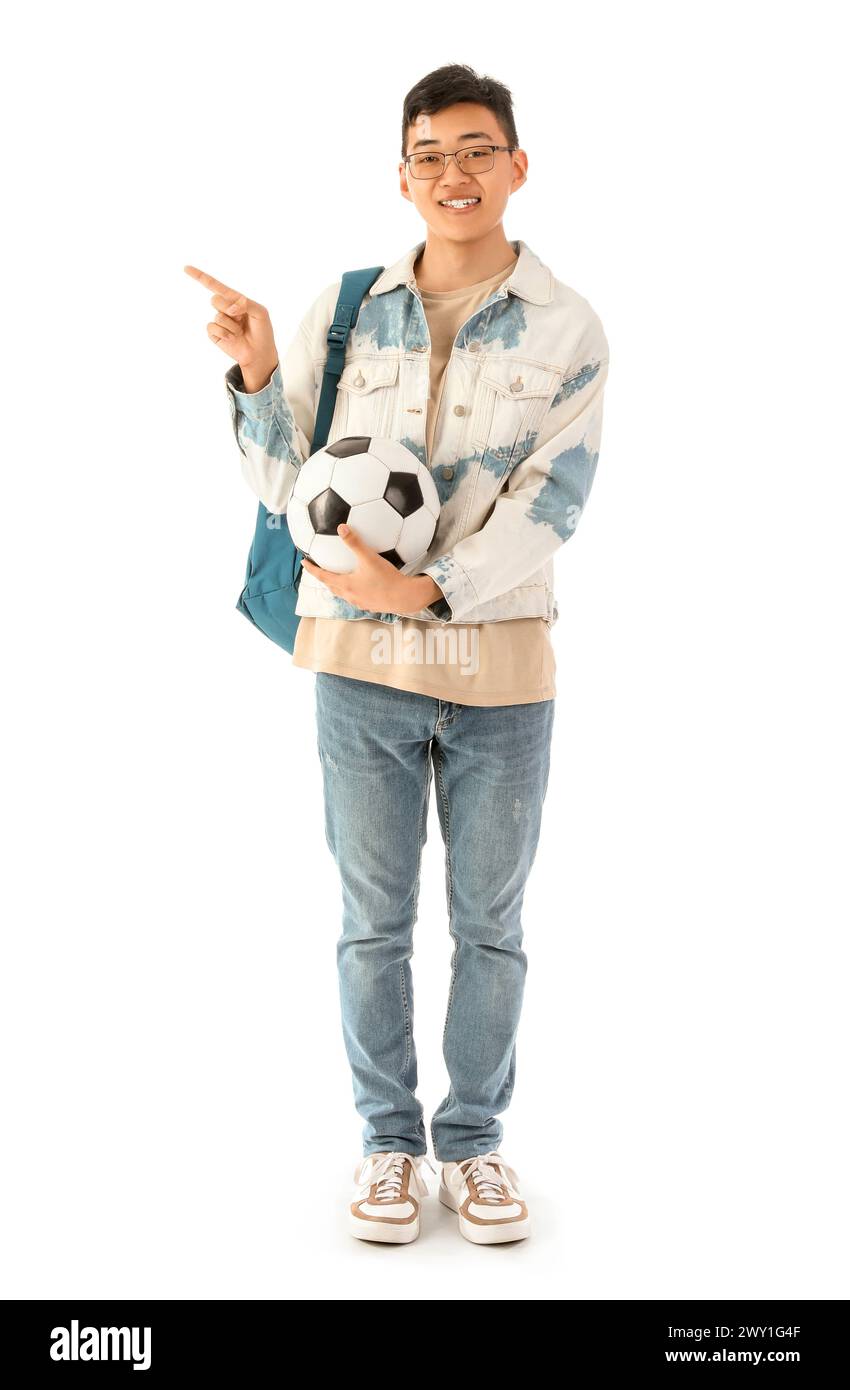Glücklicher männlicher asiatischer Student mit Rucksack und Fußball, der auf etwas auf weißem Hintergrund zeigt Stockfoto