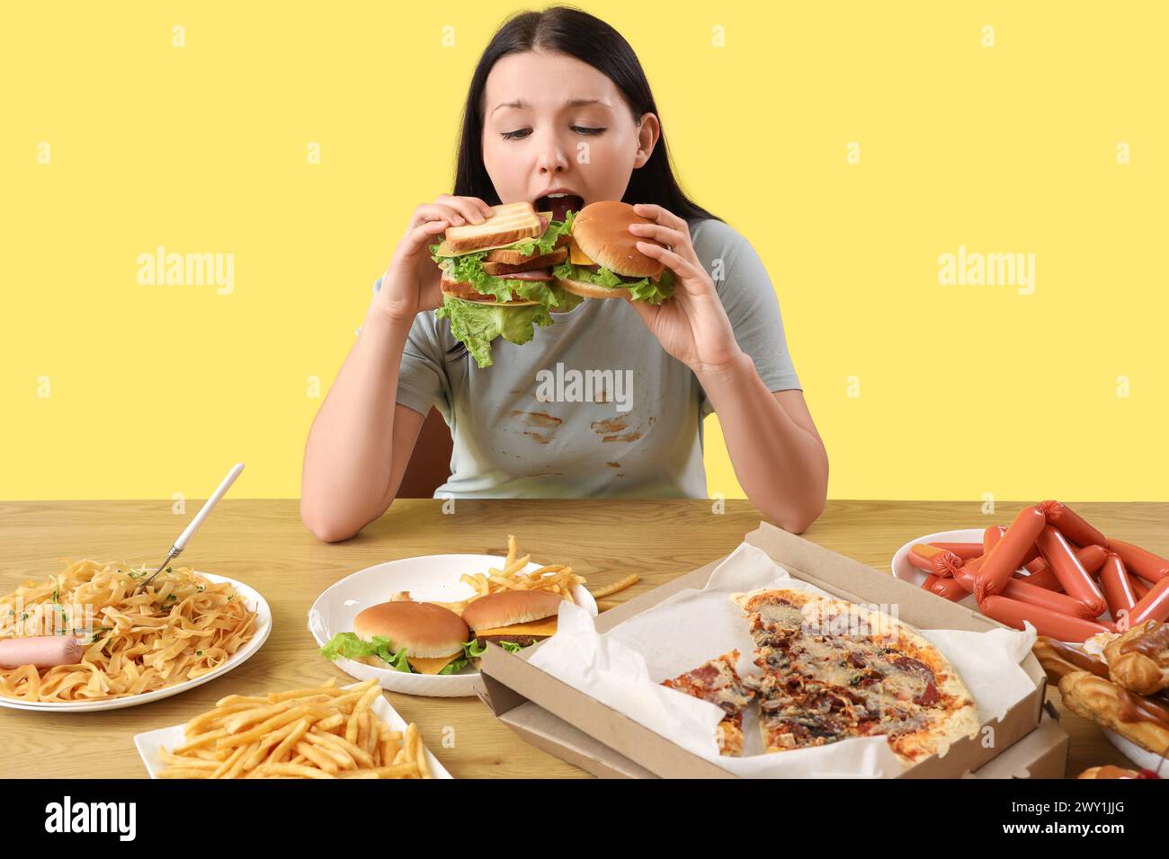 Junge Frau mit ungesundem Essen am Tisch auf gelbem Hintergrund. Das Konzept des Überessens Stockfoto
