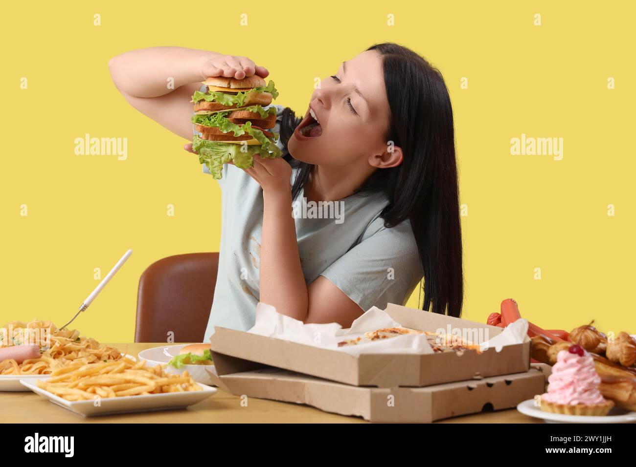 Junge Frau mit ungesundem Essen am Tisch auf gelbem Hintergrund. Das Konzept des Überessens Stockfoto