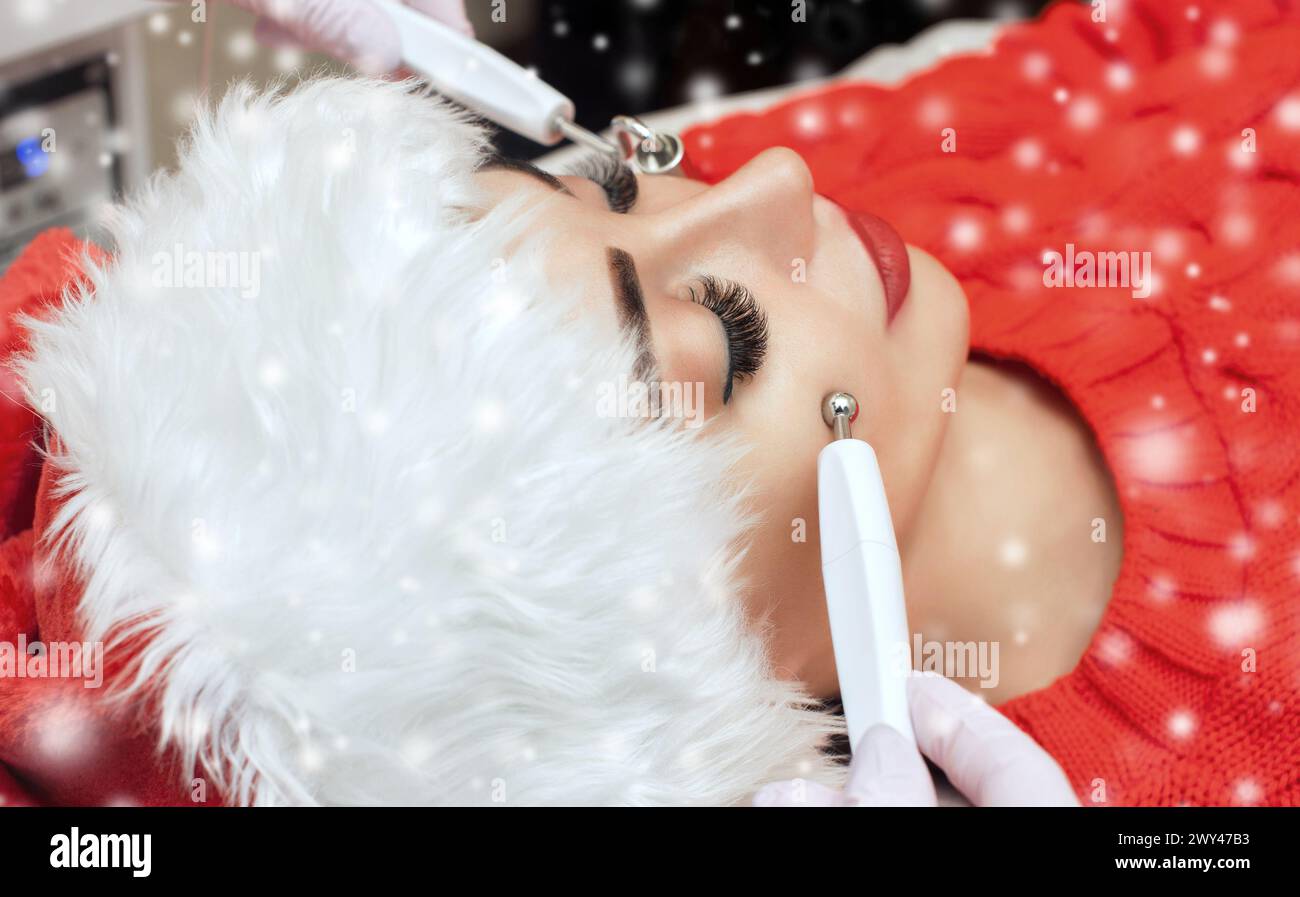 Der Kosmetologe macht das Verfahren der Mikrostromtherapie einer Frau in Santa Claus Hut auf einem Hintergrund von Schnee. Neujahrs- und Kosmetologie-Conce Stockfoto