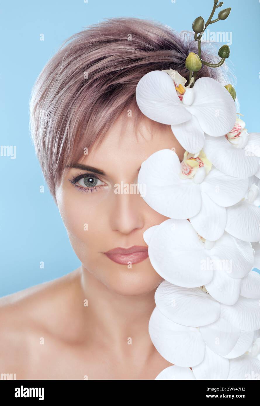 Porträt einer schönen Frau in einem Spa-Salon mit weißer Orchidee in der Hand auf blauem Hintergrund. Professionelle Hautpflege. Stockfoto