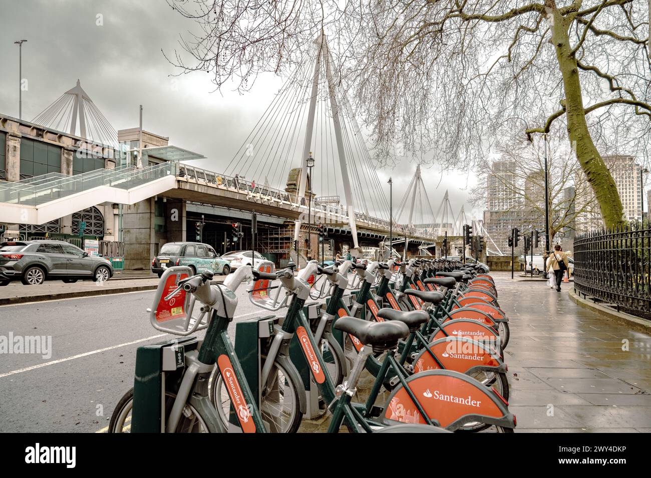 Eine Reihe von Santander E-Bikes zum Verleih in Central London. Im Hintergrund überqueren Fußgänger die Themse auf der Golden Jubilee Bridge. Stockfoto