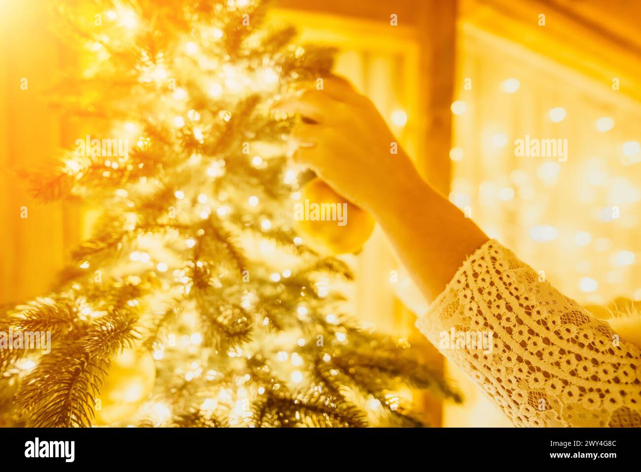 Eine Frau schmückt Einen Weihnachtsbaum mit goldenen Ornamenten und Lichtern. Der Baum ist mit Goldkugeln dekoriert und mit Lichtern beleuchtet. Stockfoto