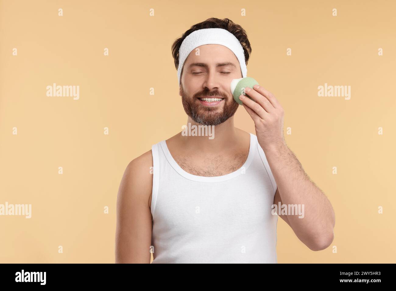 Mann mit Stirnband, der sein Gesicht mit einem Schwamm auf beigem Hintergrund wäscht Stockfoto