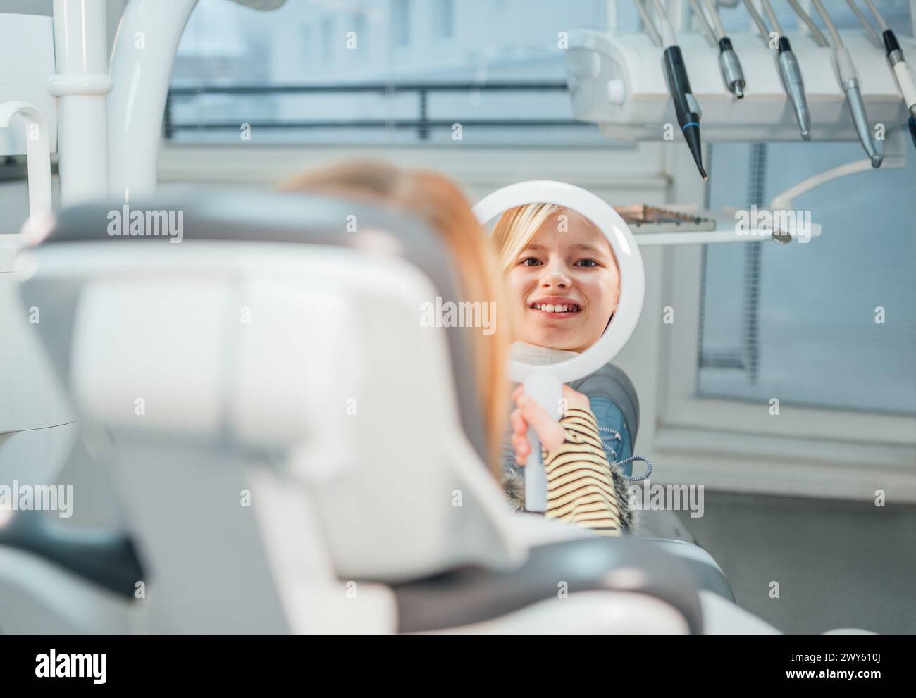 Kleines Mädchen, das im Stomatologie-Klinikstuhl sitzt und im Spiegel lächelt, der nach zahnärztlichen Eingriffen ihre Zähne zeigt. Gesundheitsfürsorge, Kindergesundheit und ich Stockfoto