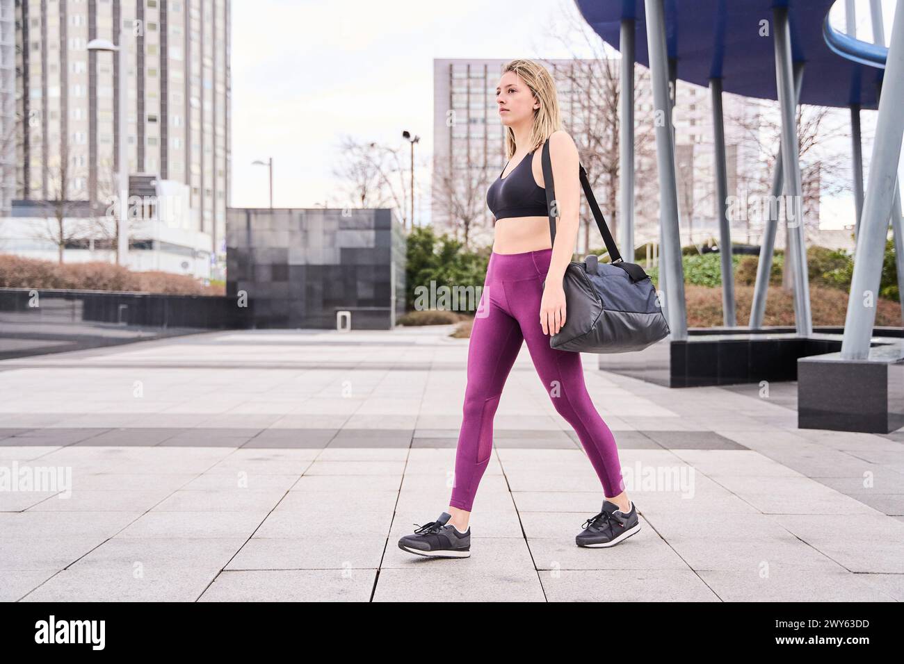 Blonde Frau in Sportbekleidung, die mit einem Rucksack durch die Stadt läuft, auf dem Weg zum Fitnessstudio Stockfoto
