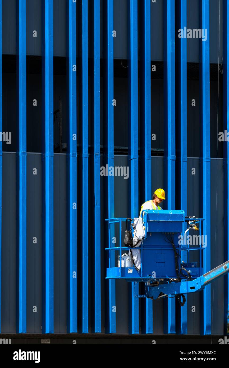 Ein Arbeiter, der eine hydraulische Hubplattform für Wartungs- und Bauarbeiten verwendet, um Verkleidungsabschnitte an der Außenseite eines Gebäudes aufrechtzuerhalten. Stockfoto