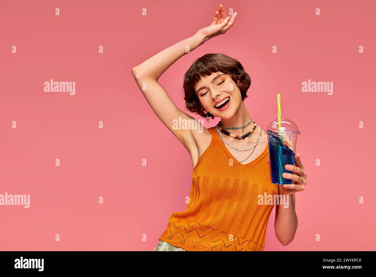 Aufgeregte junge Frau mit kurzen brünetten Haaren, die erfrischendes Sommergetränk über dem Kopf auf rosa hält Stockfoto