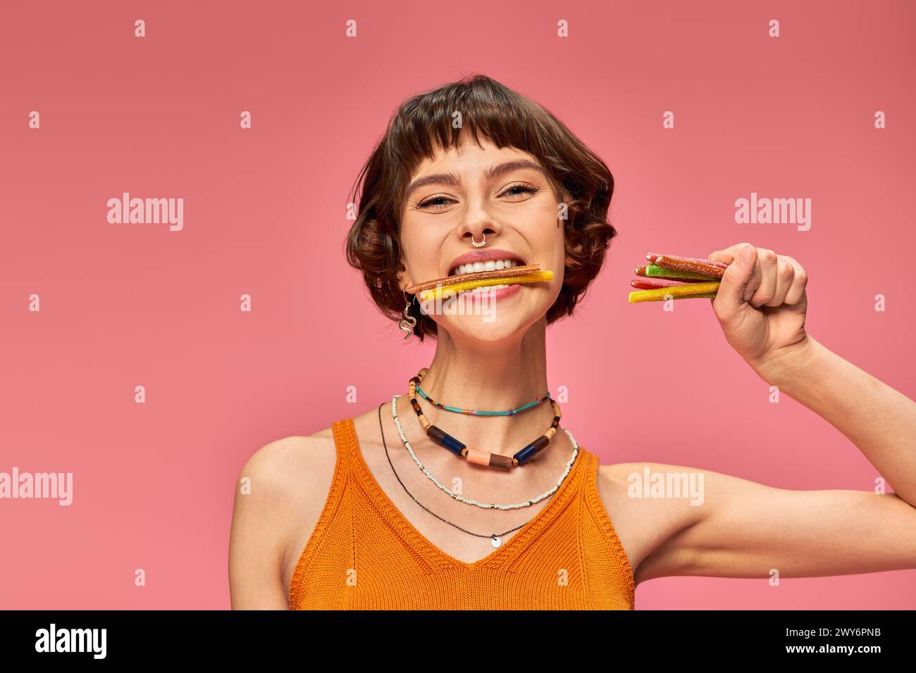 Glückliche junge Frau in ihren 20ern mit süßen und sauren Süßigkeiten in Händen und Zähnen auf rosa Hintergrund Stockfoto