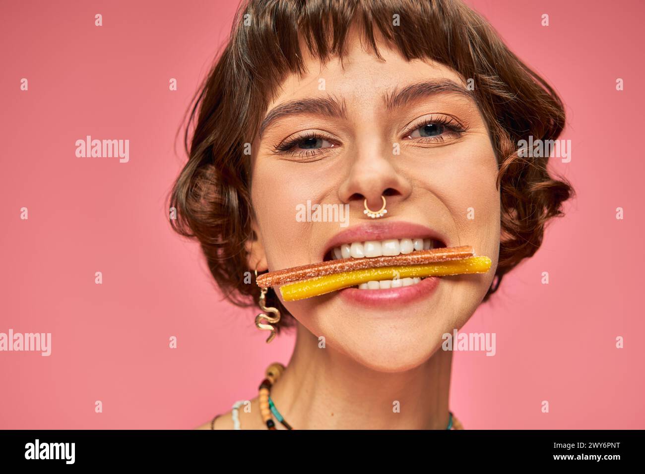 Nahaufnahme einer glücklichen Frau in ihren 20ern mit süßen und sauren Süßigkeiten in ihren weißen Zähnen auf rosa Hintergrund Stockfoto