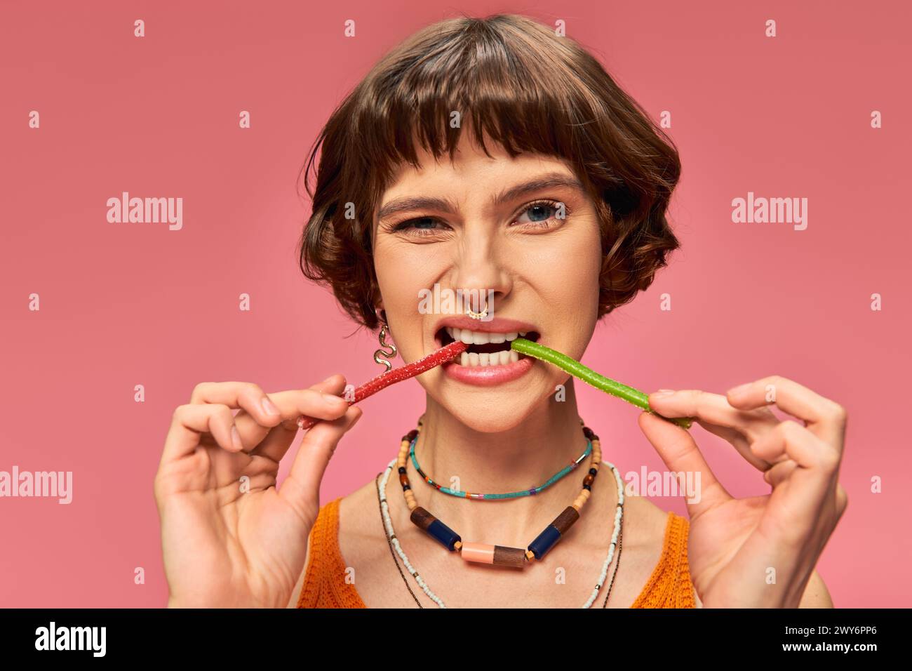 Lustiges durchbohrtes Mädchen in 20ern beißt zwei verschiedene Geschmacksrichtungen süßer und saurer Süßigkeiten vor rosa Hintergrund Stockfoto
