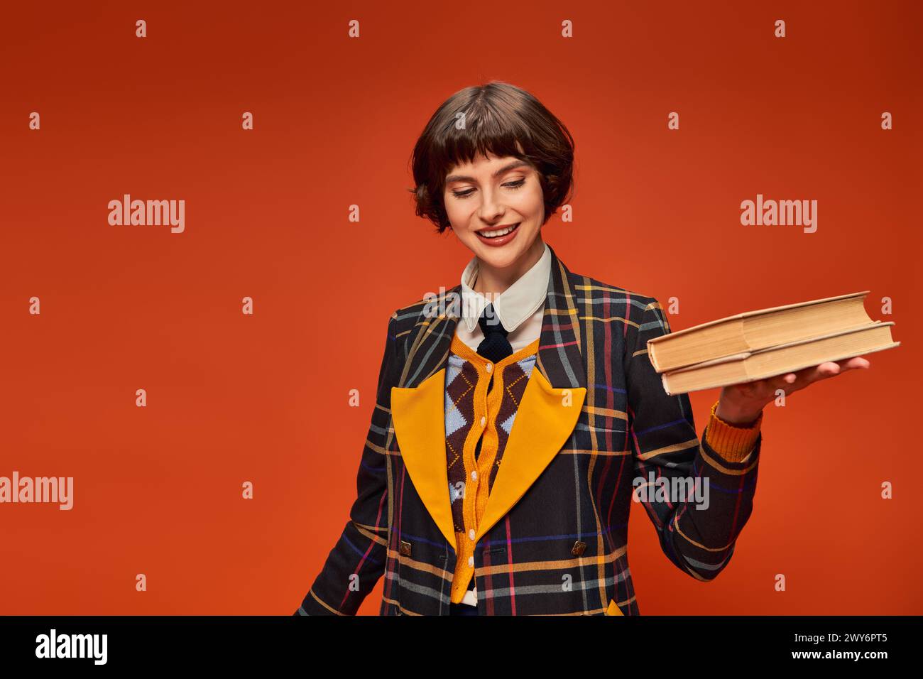 Glückliches College-Mädchen in Uniform mit einem Stapel Bücher in der Hand auf orangem Hintergrund, Wissen Stockfoto