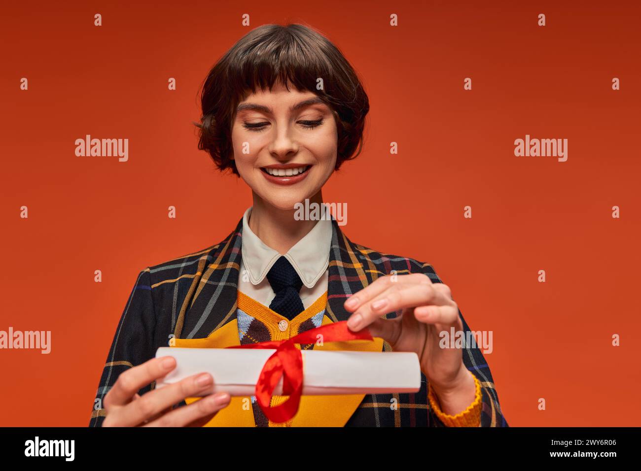 Porträt eines glücklichen College-Mädchens in karierter Uniform mit Blick auf das Abschlusszeugnis vor orangem Hintergrund Stockfoto