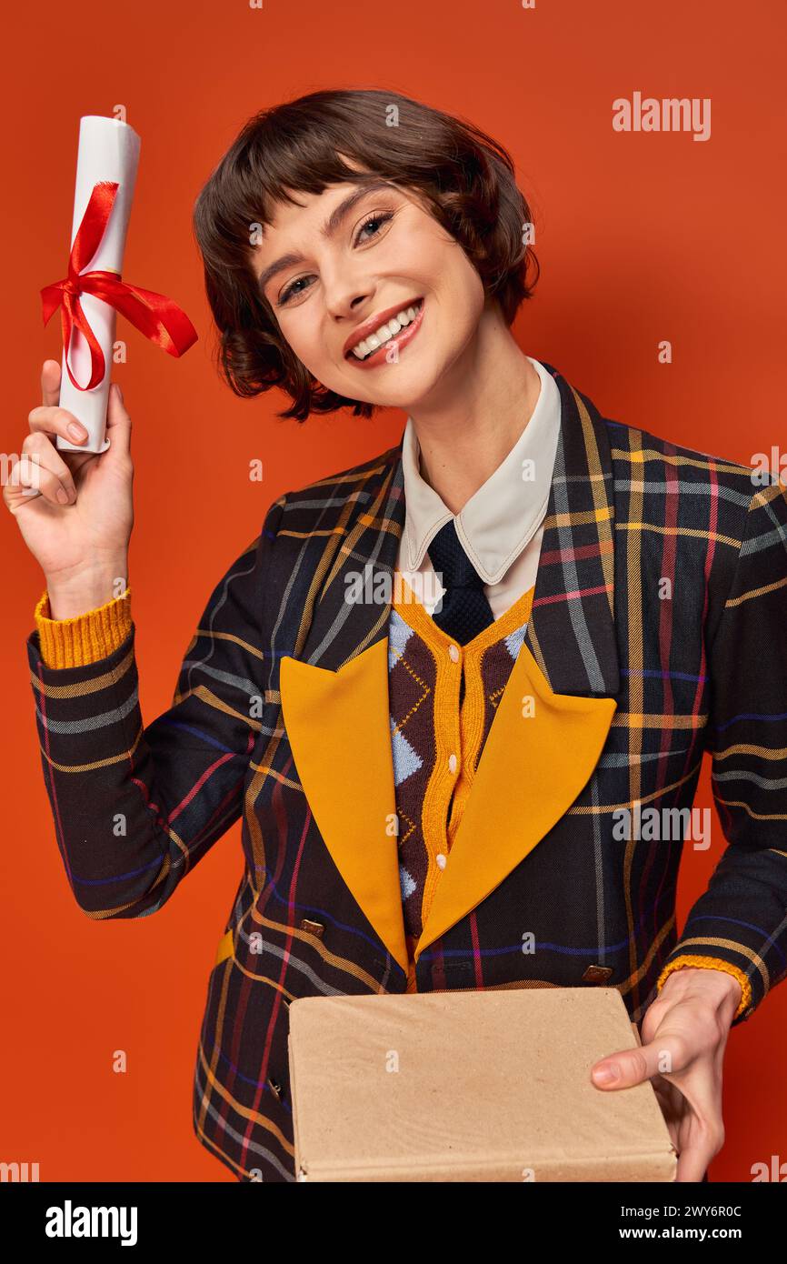 Porträt eines aufgeregten College-Mädchens in karierter Uniform mit Büchern und Diplom vor orangem Hintergrund Stockfoto
