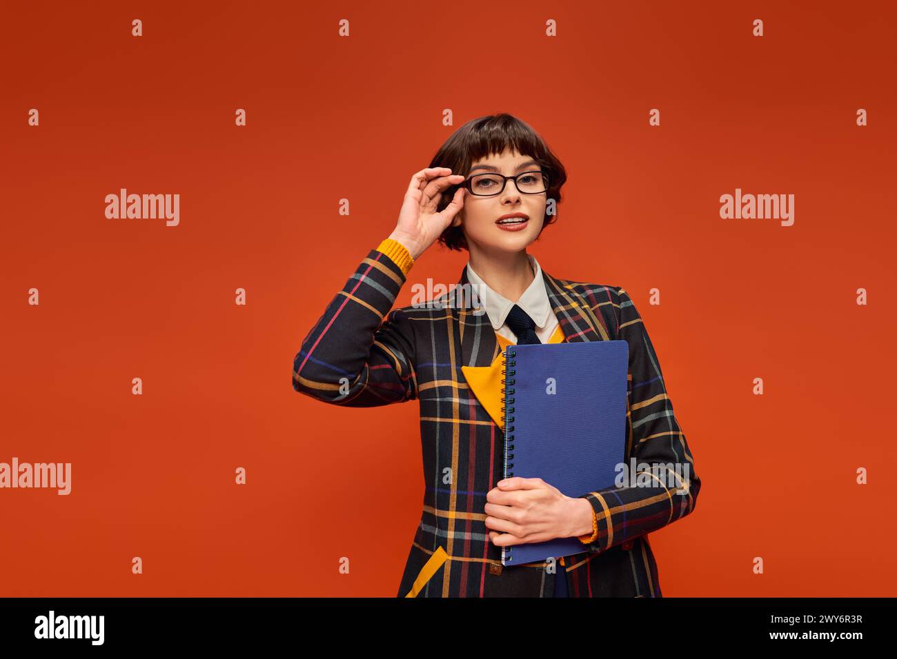 Nachdenkliche Studentin in Uniform, die ihre Brille anpasst und Notizbuch auf orangefarbenem Hintergrund hält Stockfoto