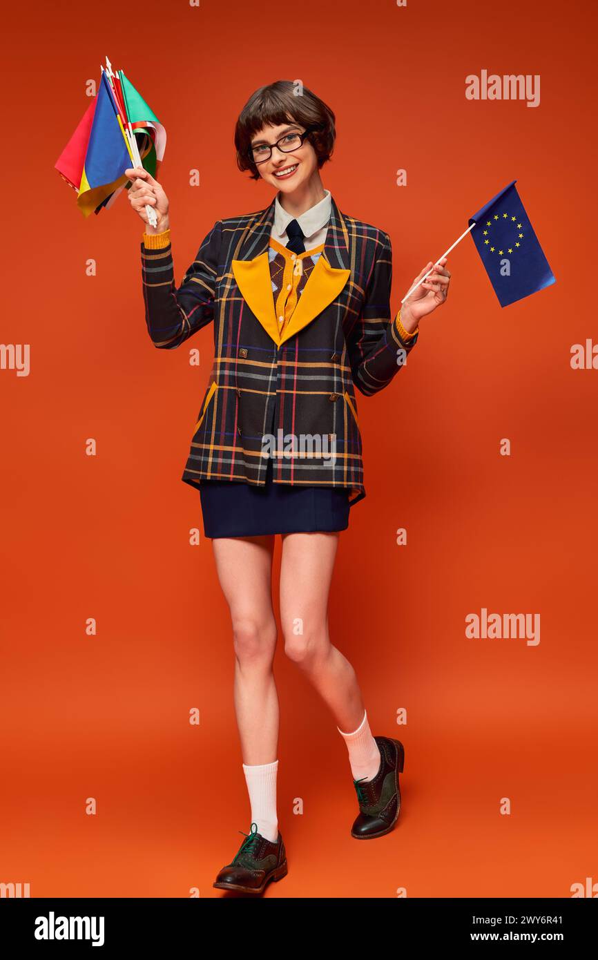 Glückliches junges College-Mädchen in Uniform und Brille, die EU und verschiedene Fahnen auf orangem Hintergrund hält Stockfoto