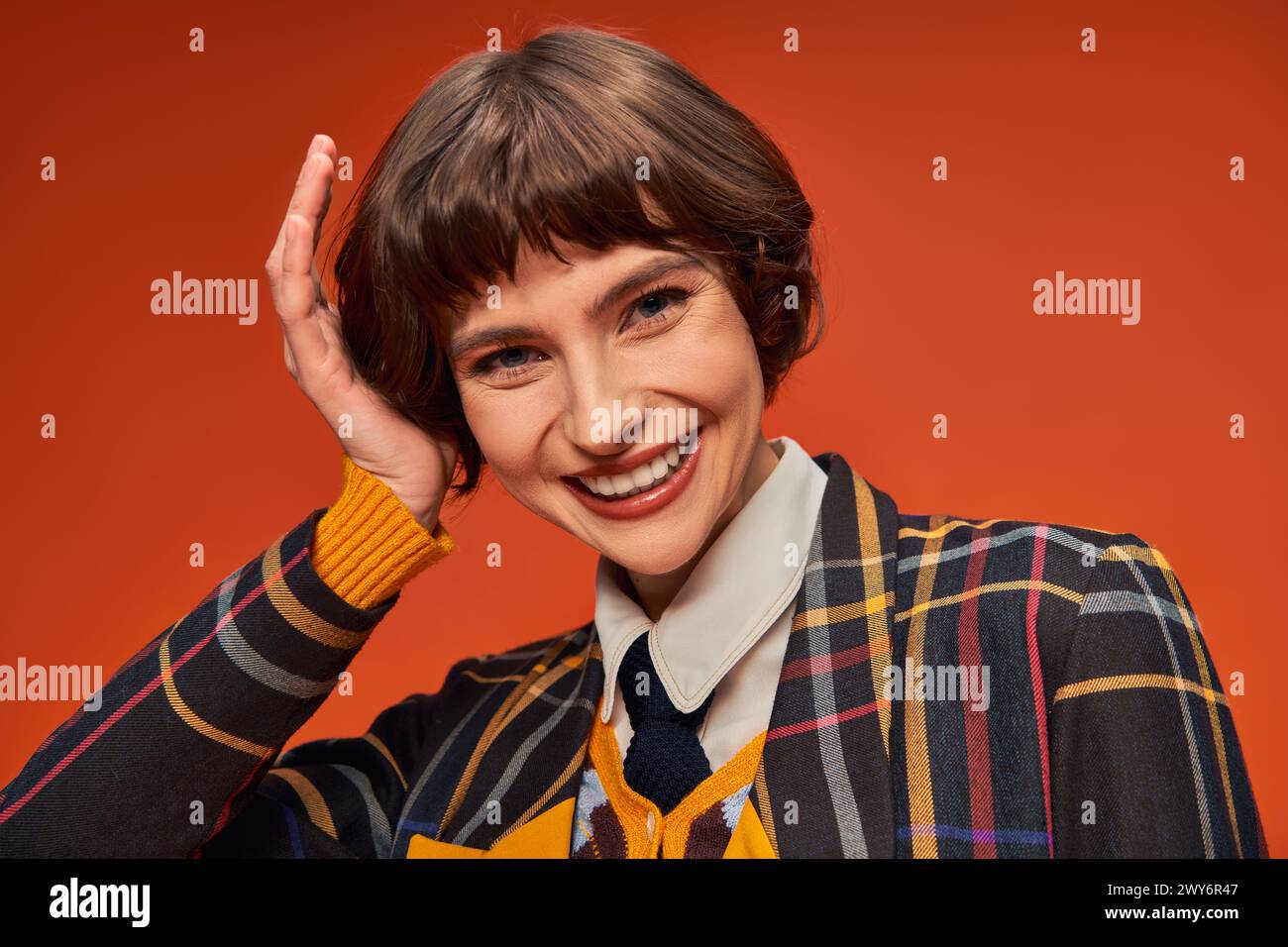 Glückliches College-Mädchen mit kurzen Haaren posiert in ihrer Uniform auf orangem Hintergrund, High School Stockfoto