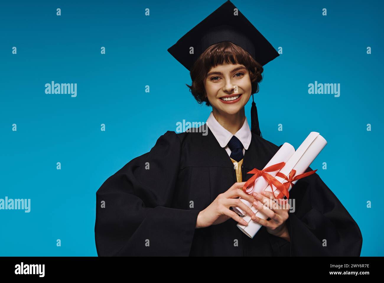 Glückliches College-Mädchen mit Abschluss im Kleid und akademischer Mütze, das Diplome mit Stolz auf Blau hält Stockfoto