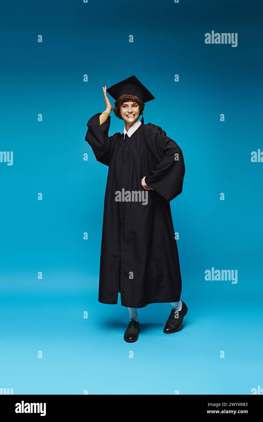 Aufgeregtes College-Mädchen in Kleid und Mütze lächelt auf blauem Hintergrund, Leistung Stockfoto