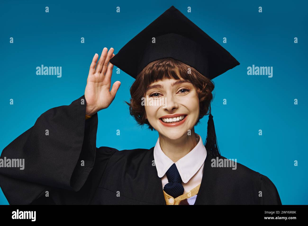 Junge und fröhliche Graduiertenkolleg-Mädchen in akademischer Mütze, grüßt mit Hand, blauer Hintergrund Stockfoto