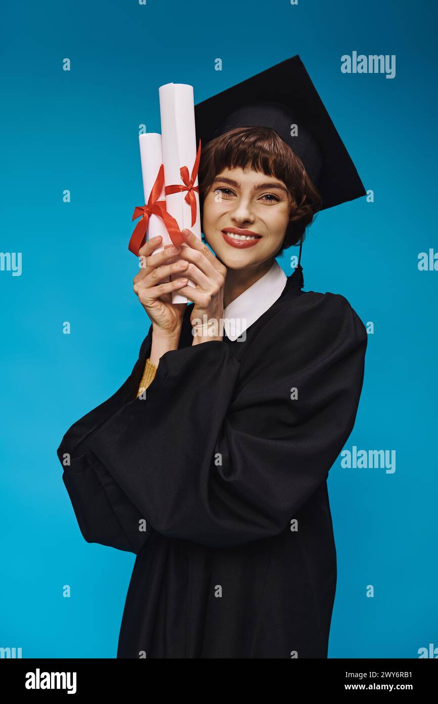 Eifriges College-Mädchen im Kleid und akademischer Mütze, das Diplome mit Stolz und blauem Hintergrund hält Stockfoto